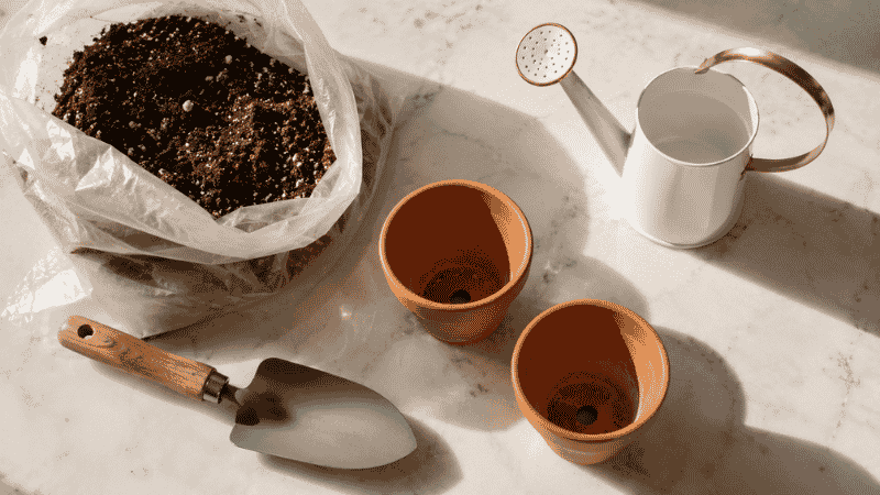 Beginner gardening essentials laid flat: a small trowel, bag of potting mix, watering can, and two pots on a white surface
