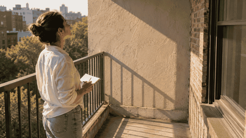 A person standing on an empty apartment balcony observing sunlight patterns — spending one day watching your balcony's light is the most important preparation step before starting a balcony garden